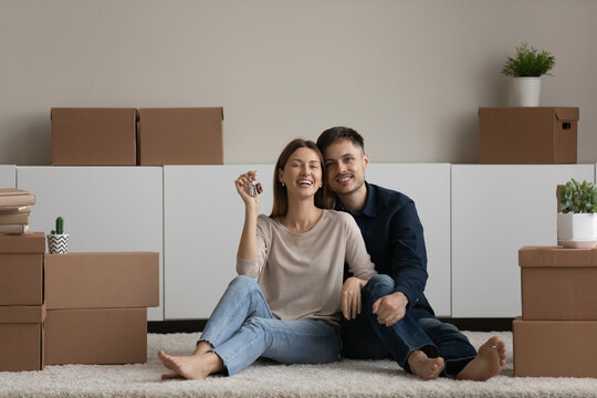 Portrait happy couple demonstrating showing keys, looking at camera, sitting on floor with cardboard boxes in new home, enjoying relocation, smiling woman and man hugging, celebrating moving day