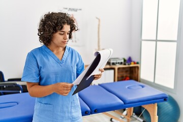 Young hispanic woman wearing physio therapist uniform reading paperwork at clinic
