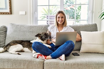Young caucasian girl smiling happy sitting on the sofa with dog using laptop at home.
