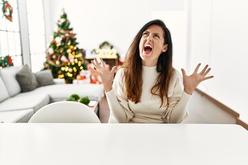 Obraz na płótnie Canvas Beautiful caucasian woman sitting on the table by christmas tree crazy and mad shouting and yelling with aggressive expression and arms raised. frustration concept.