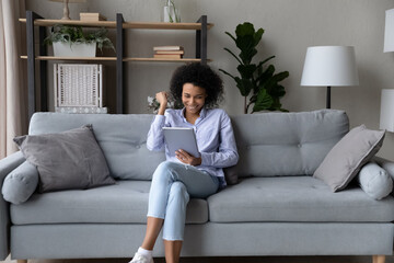 Happy African American woman showing yes gesture, looking at tablet screen, sitting on couch,...
