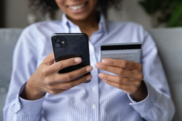 Cropped close up of African American woman paying credit card online, holding smartphone in hands, happy young female entering information, browsing internet banking service, purchasing goods