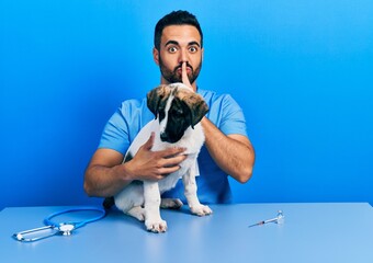 Handsome hispanic veterinary man with beard checking dog health asking to be quiet with finger on lips. silence and secret concept.
