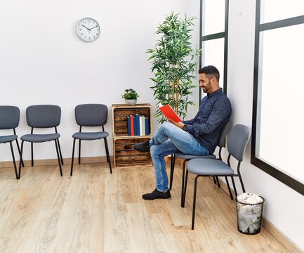 Young hispanic man reading book sitting on chair at waiting room
