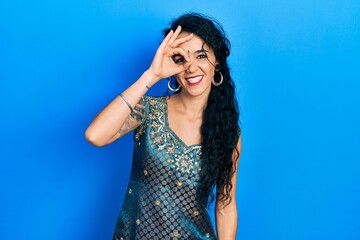 Young woman wearing bindi and traditional kurta dress doing ok gesture with hand smiling, eye looking through fingers with happy face.