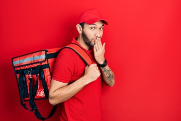 Hispanic man with beard wearing delivery package backpack covering mouth with hand, shocked and...