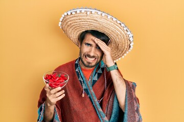 Young hispanic man wearing mexican hat holding chili stressed and frustrated with hand on head, surprised and angry face