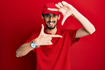 Young hispanic man wearing delivery uniform and cap smiling making frame with hands and fingers with happy face. creativity and photography concept.