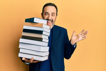 Middle age hispanic man holding a pile of books celebrating achievement with happy smile and winner expression with raised hand