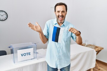 Middle age man with beard at political campaign election holding estonia flag celebrating achievement with happy smile and winner expression with raised hand