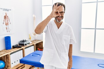 Middle age man with beard working at pain recovery clinic doing ok gesture with hand smiling, eye looking through fingers with happy face.