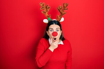 Young hispanic woman wearing deer christmas hat and red nose looking fascinated with disbelief, surprise and amazed expression with hands on chin