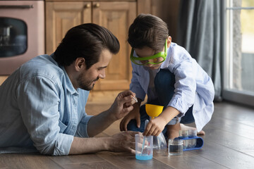Funny learning. Caring millennial father preteen son do simple chemical experiments using chemistry set for kids. School age boy study experimental science in education game playing on floor with dad