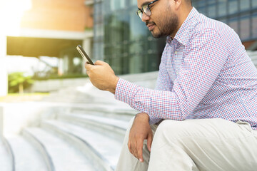 Bearded businessman smiling wearing glasses using modern smartphone device typing text message in social media while sitting on stairs outdoor. Happy male spending free time in break of working.