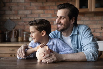 Investment in happy future. Young dad little tween boy sit at table hug hold piggy bank look at...