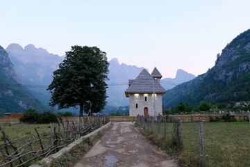 Scenery of famous mountain village Theth in Theth Valley - stone church next to dirt road with wooden fence, on background the mountains of Albanian Alps