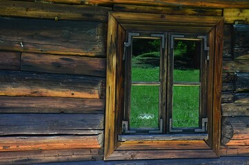 Obraz na płótnie Canvas Historical small window frame on small wooden cottage from outdoor museum in Orava region, northern Slovakia with reflection of summer green grassy meadow, sunlit by daylight sunshine. 