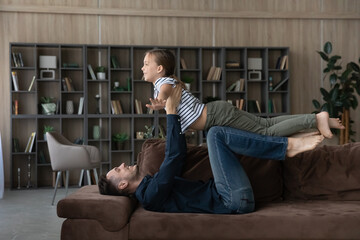 Smiling loving father lifting little daughter pretending flying on couch in living room, playing active funny game, smiling 9s girl child and dad spending leisure time together, having fun at home