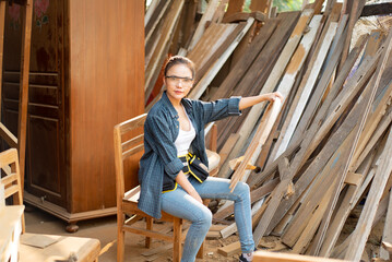Asian female carpenter in blue shirt, jean, tool belt and safety goggles sitting on chair with one hand holding the broken backrest of chair put on her leg while looking at camera in a wood factory.