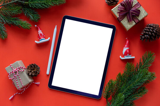 close up top view mockup of white blank digital tablet screen and stylus pen with Santa claus,pine,cone and gift on red  background for winter season collection design concept