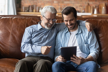 Tech and diverse generations. Elderly man in glasses use electronic tablet computer together with grownup son. Young man share video photo on digital device watch movie on screen with smiling old dad