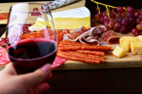 In Focus On A Wooden Board, Sliced Lard And Fuet, Cabanos Sticks, Grapes, Brie Cheese, Diced Parmesan And Prosciutto Ham. Shallow Depth Of Field. In The Foreground, Out Of Focus A Glass Of Wine.