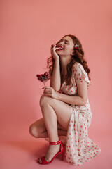 Lovely caucasian young woman squatting broadly smiling with teeth with red lipstick. Red-haired beauty with bowl of cherries in her hands is dressed in white dress with bare leg and red shoes.