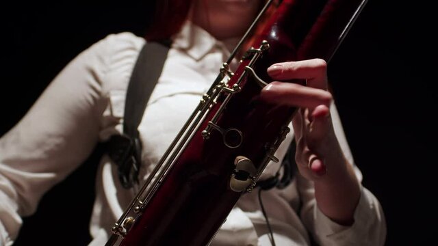Woman plays bassoon with fingers on keys, closeup, front view. Female musician plays jazz tune in studio on dark background