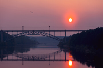 bridge over the river with two pillars in the summer at sunset. pink sky and sun reflecting in the river