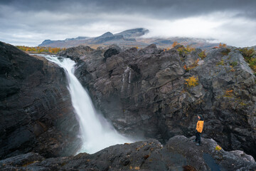 Man standing on a rock of mighty waterfall under dramatic sky. Stuor Muorkke waterfall in Stora Sjofallet national park, Sweden. Adventure in arctic wilderness