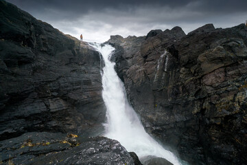 Silhouette of a man standing on the edge of mighty waterfall against dramatic sky. Stuor Muorkke waterfall in Stora Sjofallet national park, Sweden. Adventure in arctic wilderness.