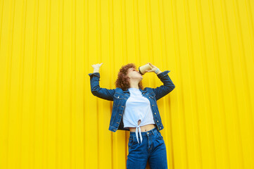 Happy curly haired woman in denim clothes drinking coffee on yellow background. Positive young woman outdoors. Smiling woman concept.