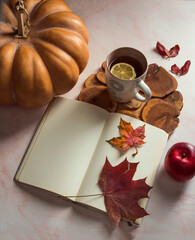 On a pink table are a mock-up of a white blank sheet in a notebook, a dry maple leaf, an orange pumpkin, a ceramic cup with tea on an old board, a ripe red apple.