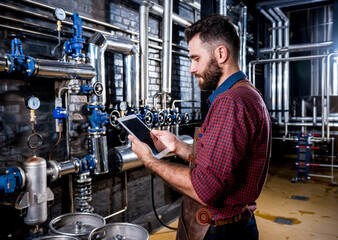 Young male brewer in leather apron supervising the process of beer fermentation at modern brewery factory
