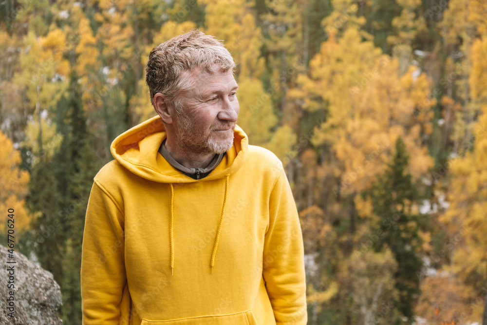 Wall mural An authentic portrait of a smiling, unshaven middle-aged traveler in a yellow hoodie on top of a mountain against the background of an autumn forest. Close-up. Selective focus. Sustainable environment - Wall murals