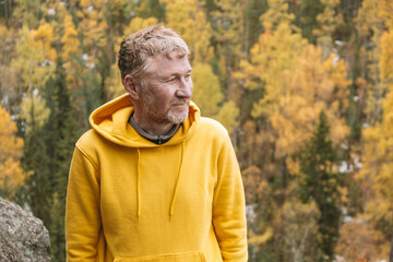 Naklejka na ściany i meble An authentic portrait of a smiling, unshaven middle-aged traveler in a yellow hoodie on top of a mountain against the background of an autumn forest. Close-up. Selective focus. Sustainable environment