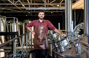 Young male brewer in leather apron supervising the process of beer fermentation at modern brewery...
