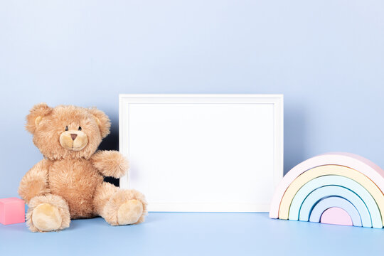 Kids toys collection. White wooden picture frame with blank mock up copy space standing next to teddy bear and toy rainbow on light blue background. Front view