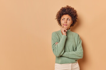 Fototapeta na wymiar Serious curly haired woman stands in thoughtful pose concentrated away ponders on something important poses against beige background with blank space for your advertising content or promotion