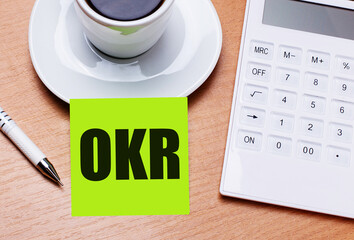 The wooden table has a white cup of coffee, a pen, a white calculator, and a green sticker with the text OKR Objective Key Results. Business concept