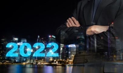 2022 new year business marketing concept. Double exposure businessman in black suit standing with his arms crossed and datum 2022 on blur night city background. Financial investment, growth planning