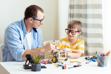 Father and son sitting by the white table and building robot at home as a school science project...
