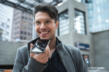 Guy wearing glasses recording voice message at his smartphone while standing at the street