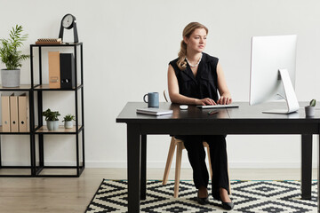 Full length portrait of blonde successful businesswoman using computer at desk in graphic black and white interior, copy space