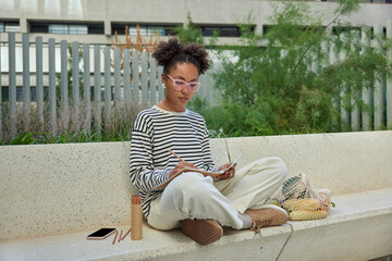Outdoor shot of teenage girl sits crossed legs makes creative drawings in notepad uses colored...