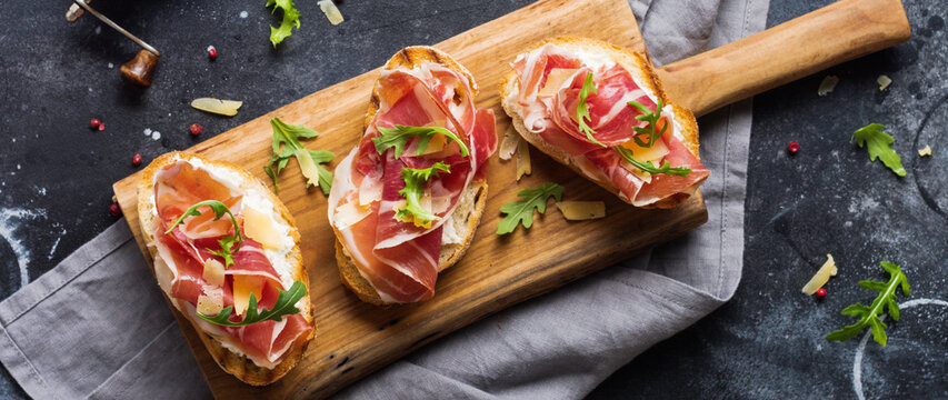 Open ham sandwiches, arugula and hard cheese, served on wooden stand with aglass of red wine on aconcrete old dark background. Rustik style. Top view