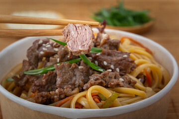 Chopsticks hold a piece of meat over a disposable paper plate of meat noodles with vegetables and herbs on a mat background. Asian street food. Close-up