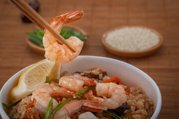 Chopsticks hold boiled shrimp over a disposable paper plate filled with rice, vegetables and herbs against the backdrop of a mat. Asian street food. Close-up