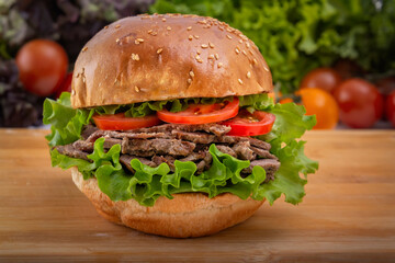 Burger with sliced lumpy meat, lettuce and tomatoes on a wooden board against a background of greens. Home cooking. Fast food. Close-up