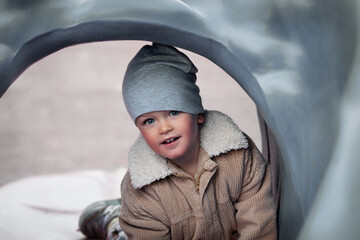 Close-up portrait of cute baby boy in beanie and jacket looking and smiling at camera. Child on gray playground outdoors